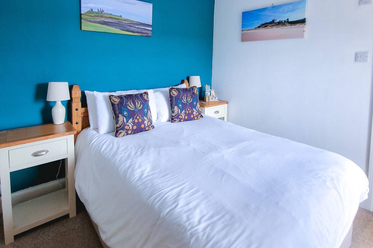 A bedroom with a large bed, two bedside tables, a nightstand, and two wall-mounted pictures.