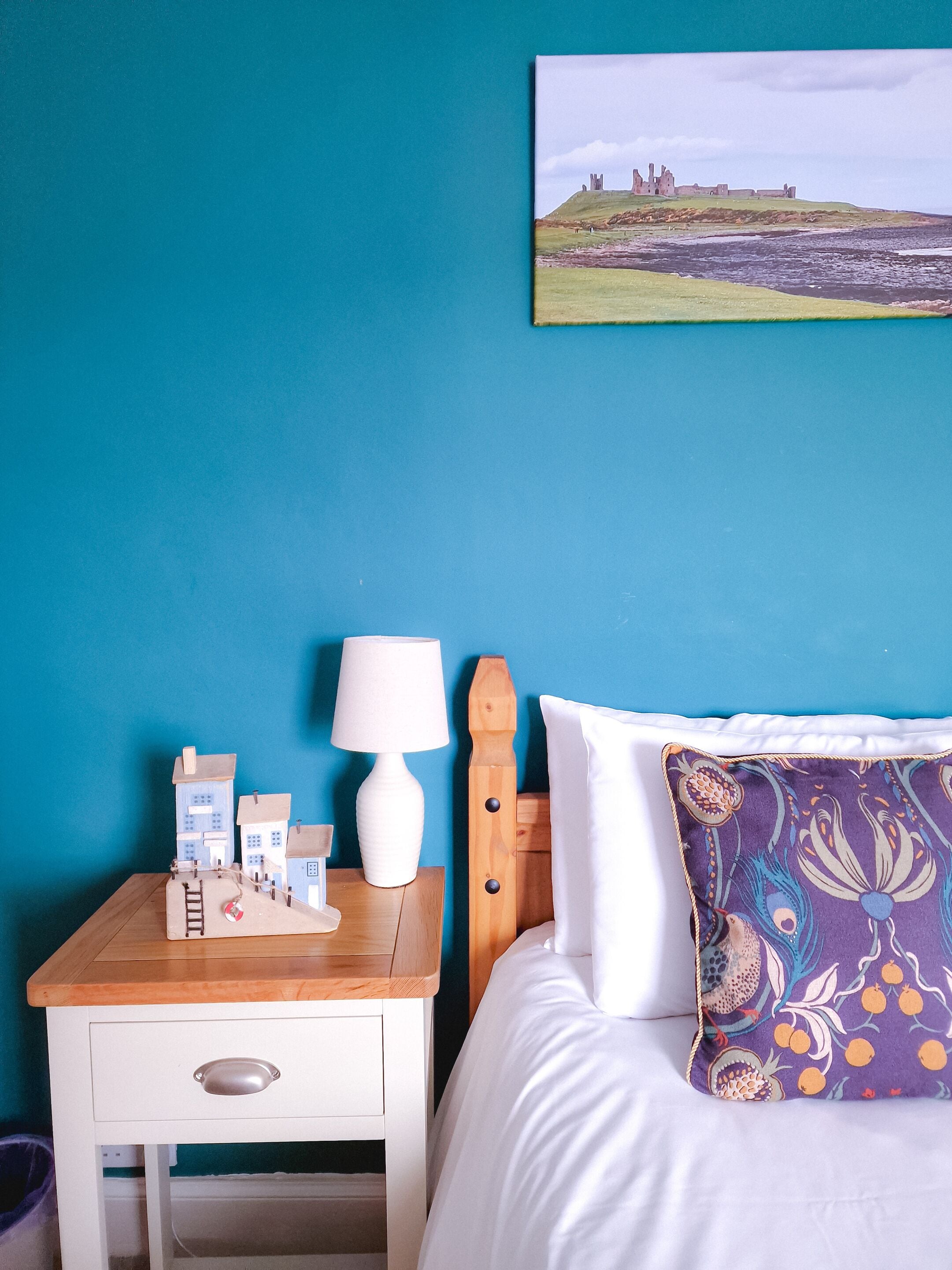 A bedroom with a bed, a nightstand with a lamp, a small table with a model, and a painting on the wall.