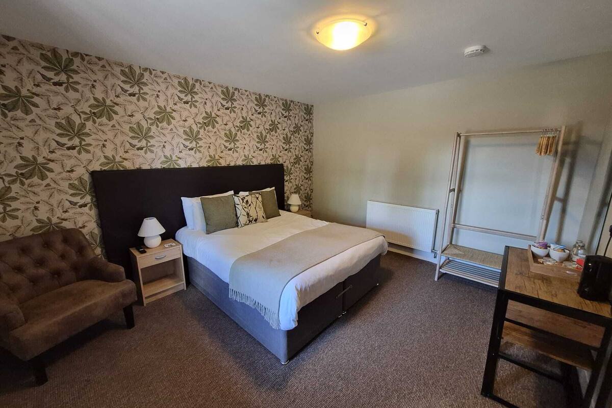 A hotel room with a bed, a chair, a desk, a radiator, and a wall with a floral pattern.