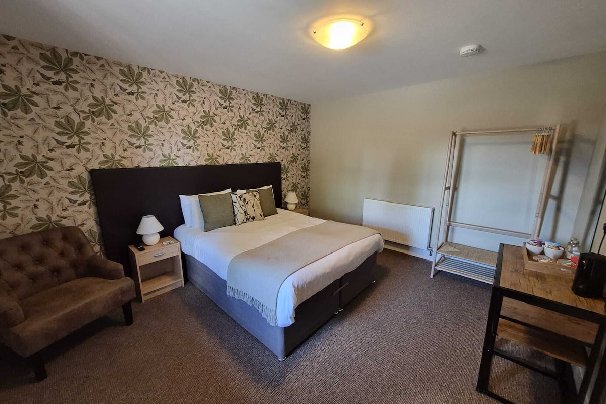 A hotel room with a bed, a chair, a desk, a radiator, and a wall with a floral pattern.