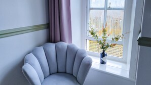 A corner room with a grey upholstered chair, a window with blinds, and a vase with a plant on the windowsill. - Red Lion Inn (Wooler)
