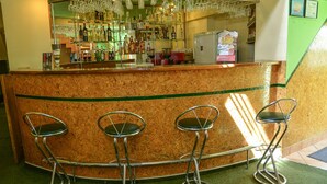 Una zona de bar con mostrador de corcho, tres taburetes y una variedad de botellas y vasos en estantes.