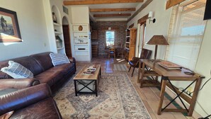 Sala de estar con un sofá de cuero marrón, una mesa de centro de madera y un tapete estampado. Hay una zona de comedor con una mesa y sillas de madera, y una cocina con pared de ladrillo y armarios de madera.