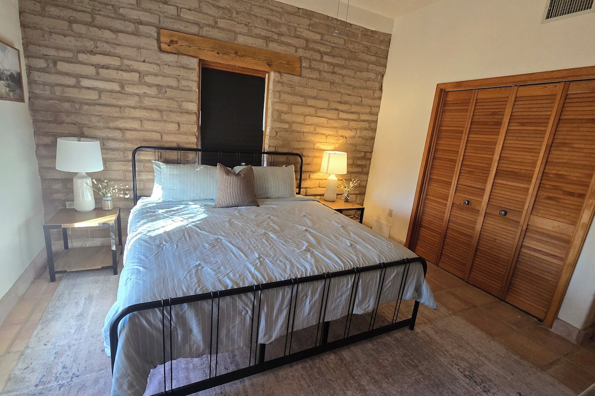 Quarto com cama, mesas de cabeceira, luminária, guarda-roupa de madeira e parede de tijolos.