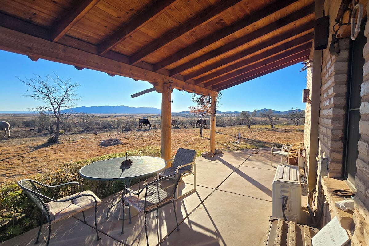 Um alpendre coberto com mesa e cadeiras, uma parede de tijolos e vista para uma paisagem árida com montanhas ao longe.