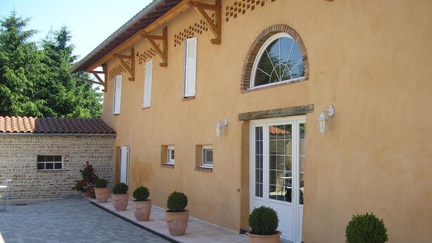 A building with a tiled roof, a stone wall, and a paved entrance area with potted plants. - Domaine de la Bellière (Villars-les-Dombes)