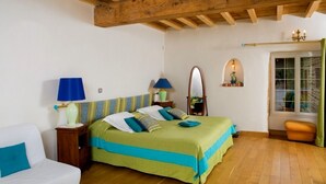 A bedroom with a wooden ceiling, a bed with green and blue bedding, a nightstand with a lamp, a mirror, and a window with curtains. - Domaine de la Bellière (Villars-les-Dombes)