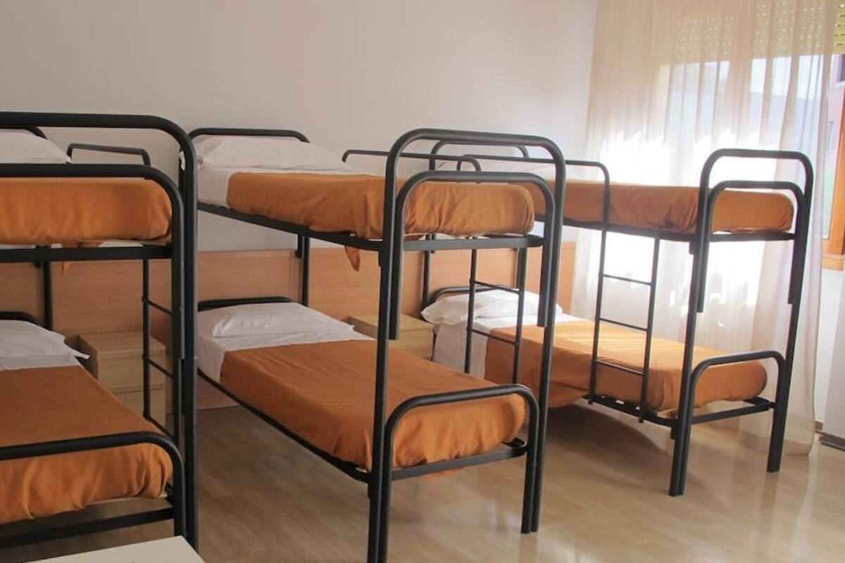 Bunk beds arranged in a dormitory-style room with a wooden floor and a window with sheer curtains.