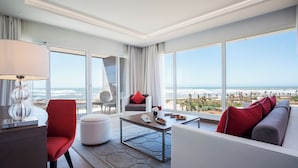 A modern hotel room with a large window offering a view of the beach and ocean. There is a red chair, a white sofa, and a small table with a lamp. - The View Bouznika (El Mansouria)