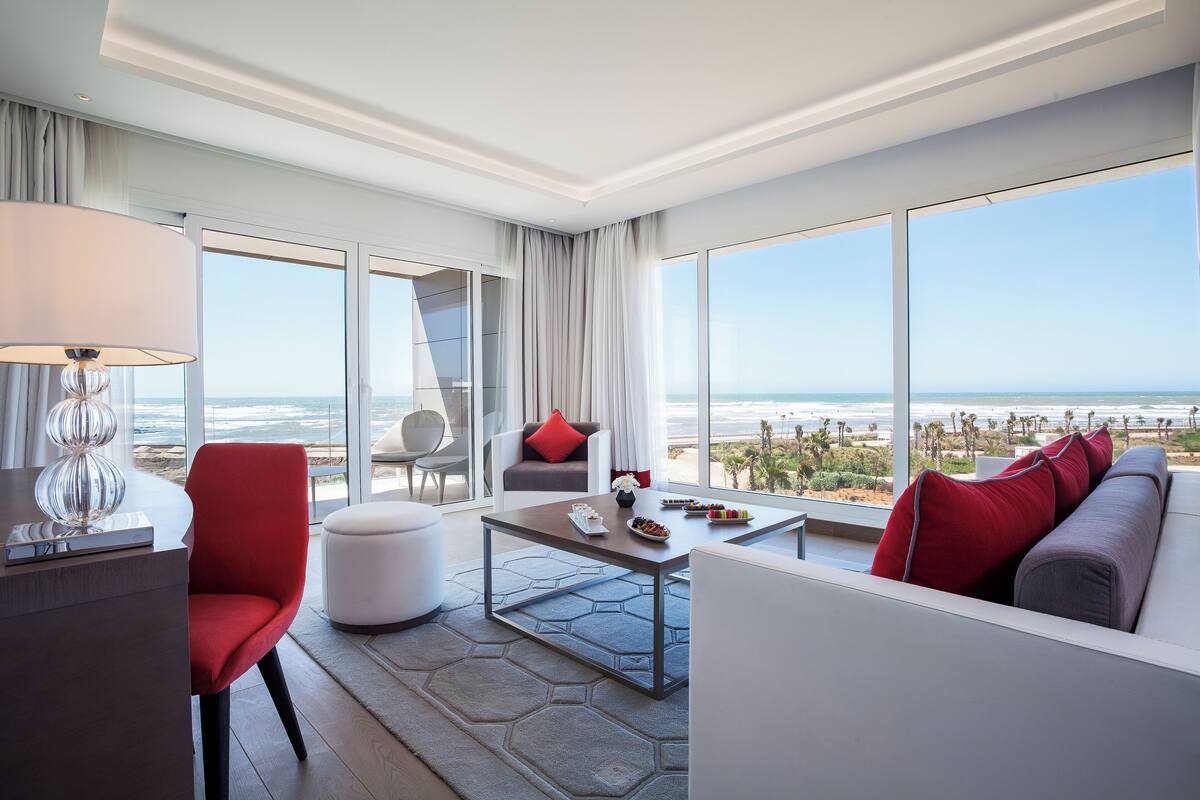 Une chambre d’hôtel moderne dotée d’une grande fenêtre donnant sur la plage et l’océan. On y trouve un fauteuil rouge, un canapé blanc et une petite table avec une lampe.