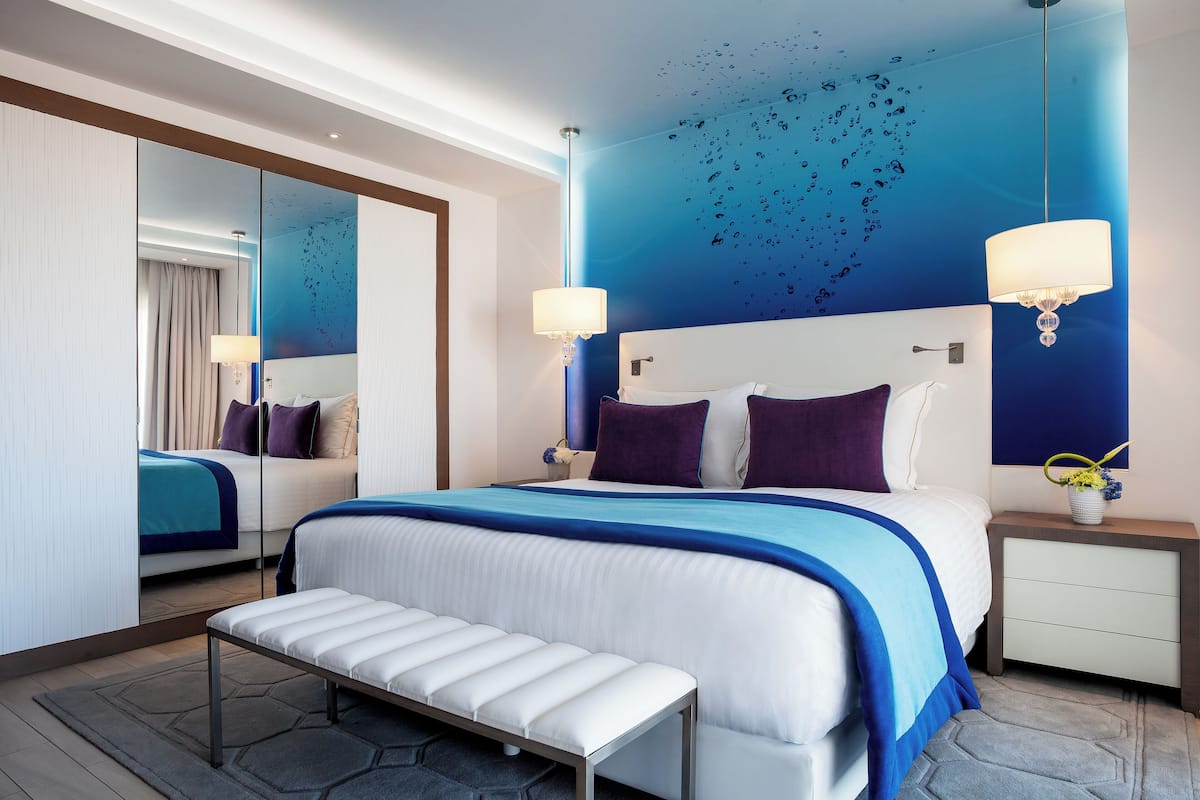 Une chambre d’hôtel moderne avec un grand lit, une banquette, une table de chevet avec une lampe et un miroir.