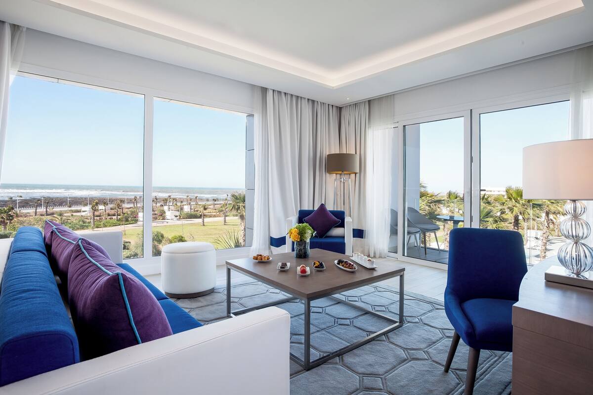 Une chambre d’hôtel moderne dotée d’une grande fenêtre offrant une vue sur l’océan et le paysage urbain. On y trouve un canapé bleu, un fauteuil blanc et une petite table agrémentée d’objets décoratifs.
