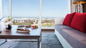 A modern room with a large window overlooking a beach, a sofa with red cushions, and a table with various snacks. - The View Bouznika (El Mansouria)