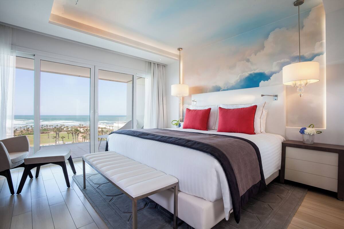 Une chambre d’hôtel moderne avec un grand lit, un banc, un bureau et une vue sur l’océan.