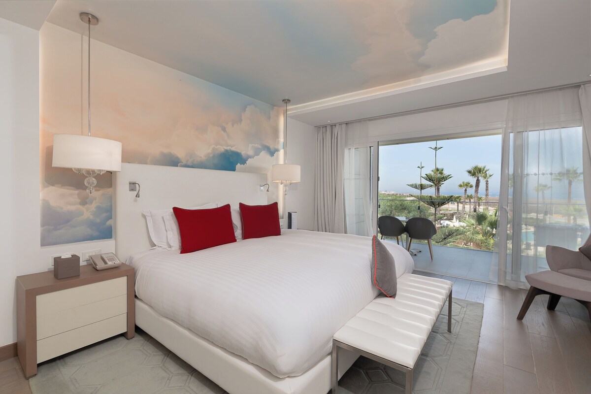 A hotel room with a large bed, a nightstand, a desk, a chair, and a view of palm trees and a cityscape through the window.