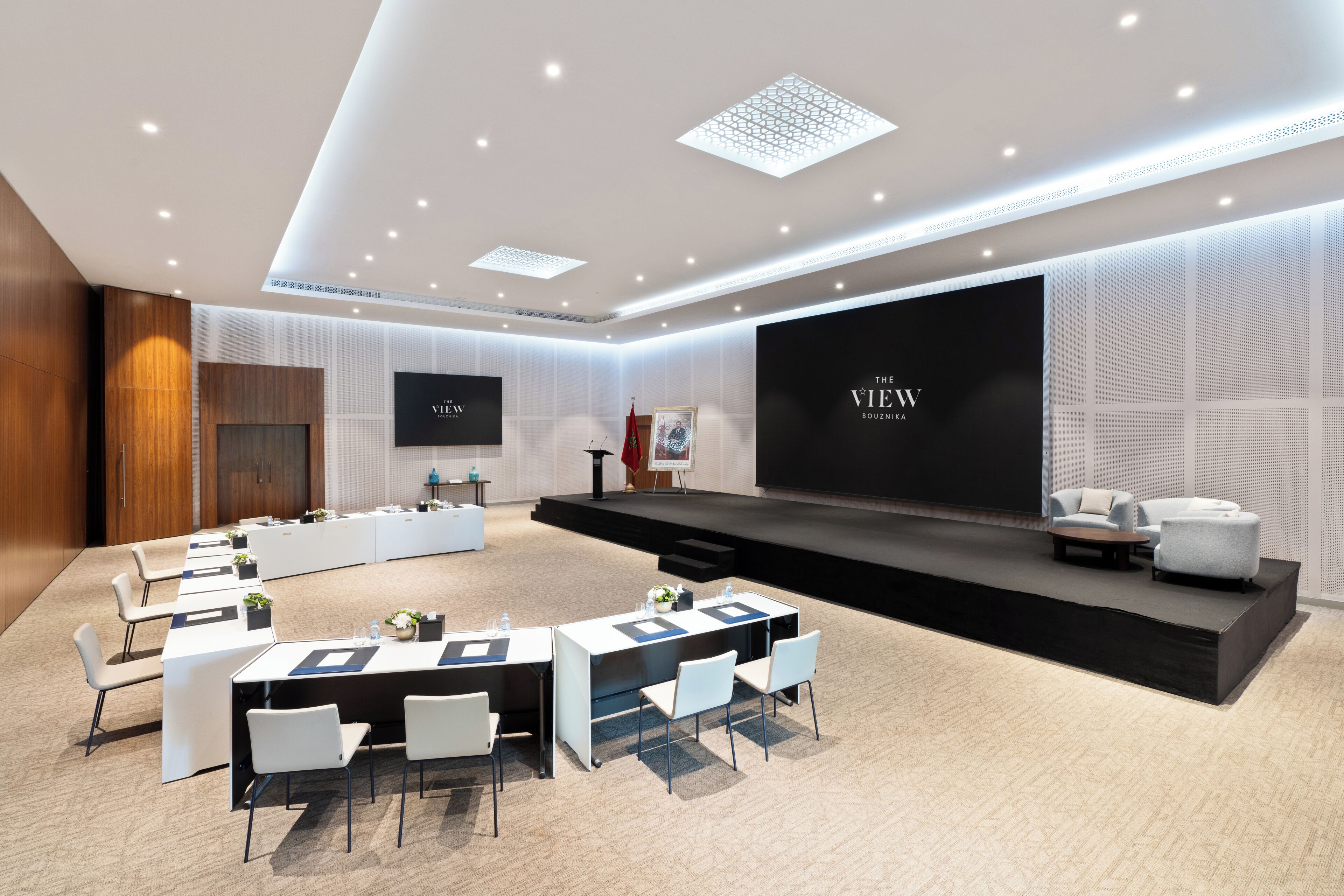 Une salle de conférence moderne équipée d’une scène, de chaises blanches, de tables et d’un grand écran diffusant l’émission « THE VIEW ».
