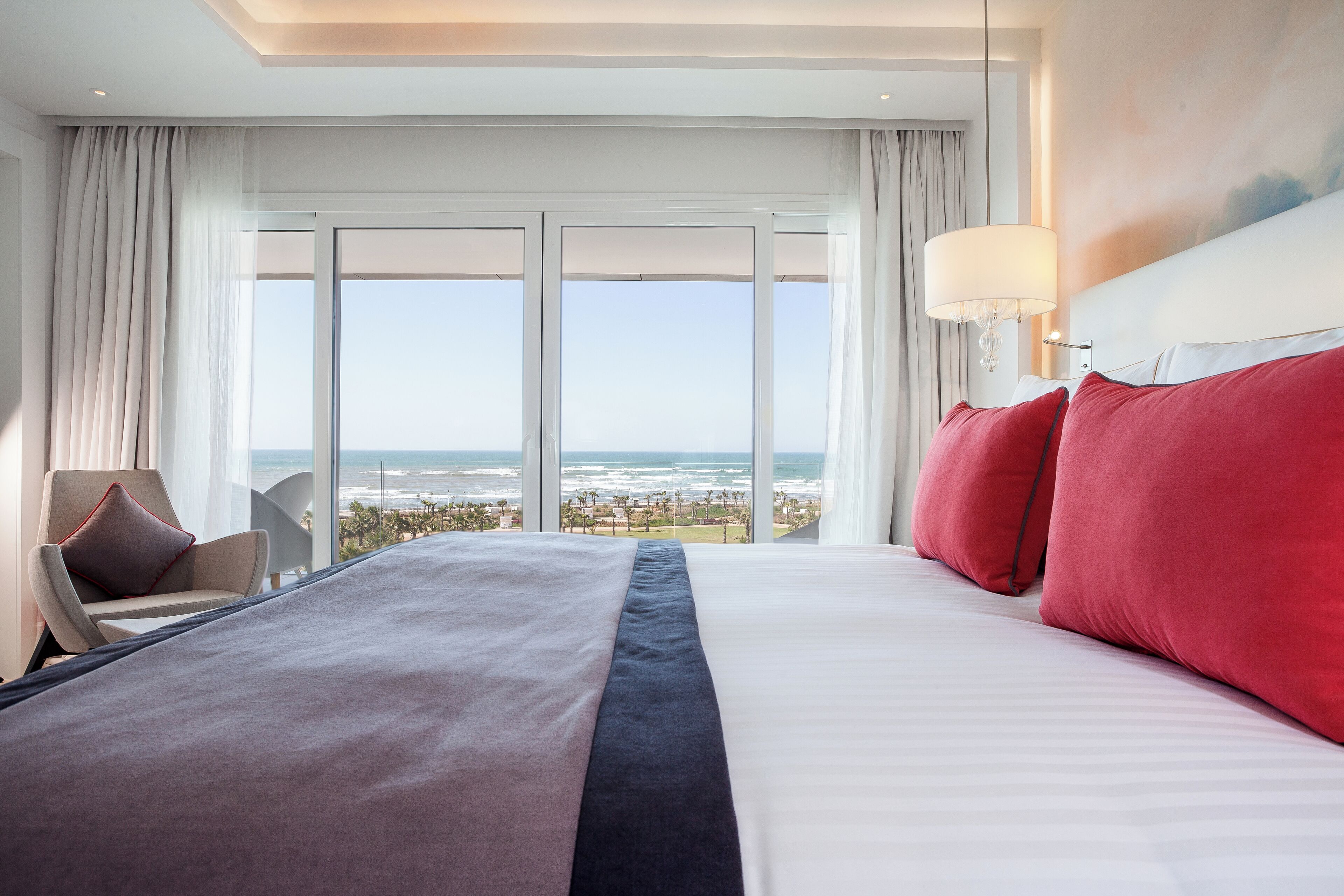 Un cuarto de hotel con una cama grande, vista al océano y una silla junto a la ventana.