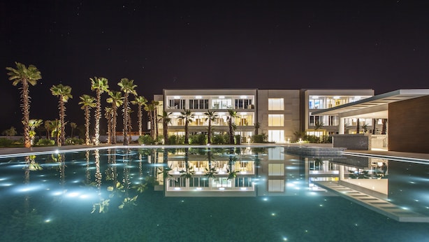 A nighttime view of a hotel with a swimming pool, palm trees, and modern architecture. - The View Bouznika (El Mansouria)
