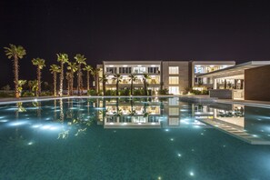 Vista nocturna de un hotel con piscina, palmeras y arquitectura moderna.