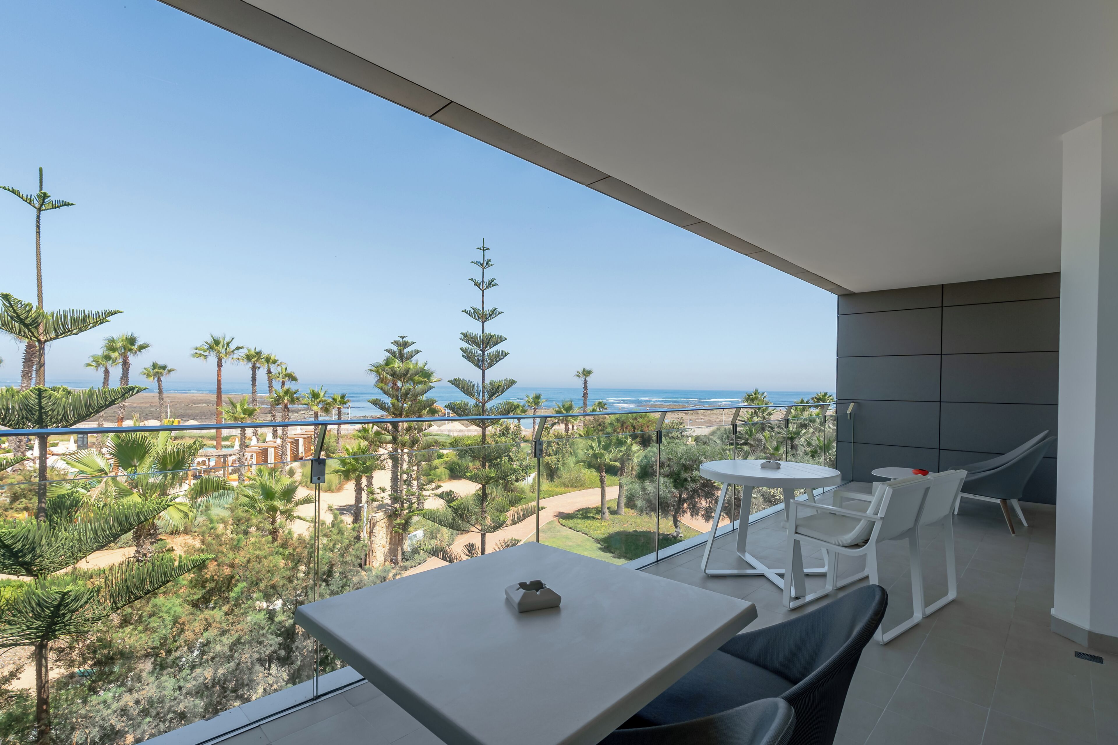 A balcony with a view of the beach and ocean. There are two white tables and chairs.