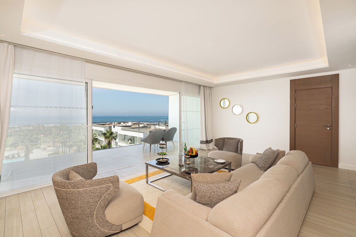 A modern living room with a large sofa, a glass coffee table, and a view of the sea.