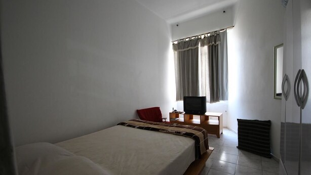 Habitación individual con una cama de madera, un escritorio pequeño con televisor y una ventana con cortinas.