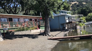 A riverside building with a dock, a large tree, and a gravel path. - Indian Beach Resort (Clearlake Oaks)