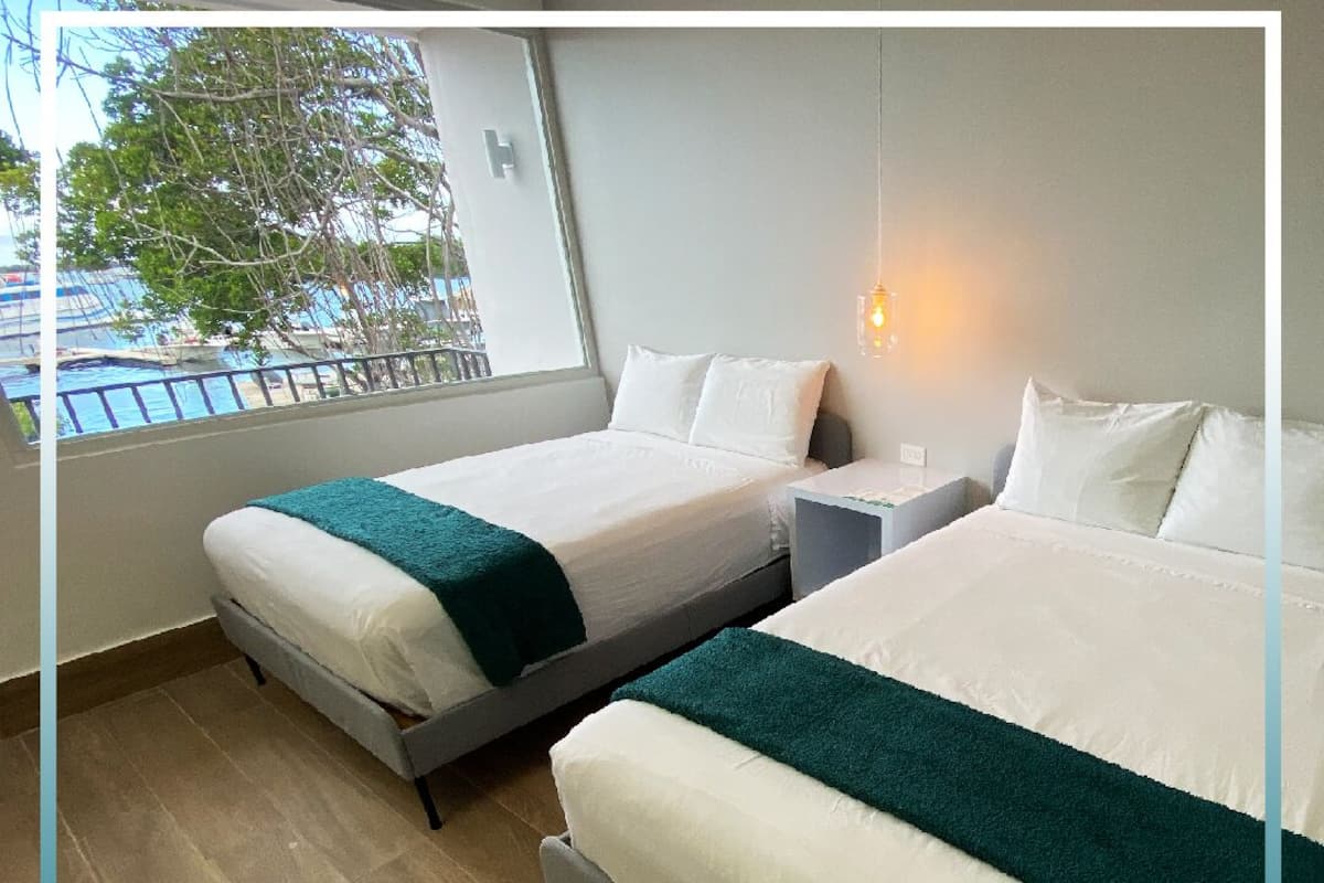 A hotel room with two beds, a nightstand, a lamp, and a window with a view of the sea.