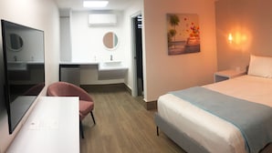 Habitación de hotel con cama, mesitas de noche, escritorio con silla, espejo, lavamanos y un cuadro en la pared.