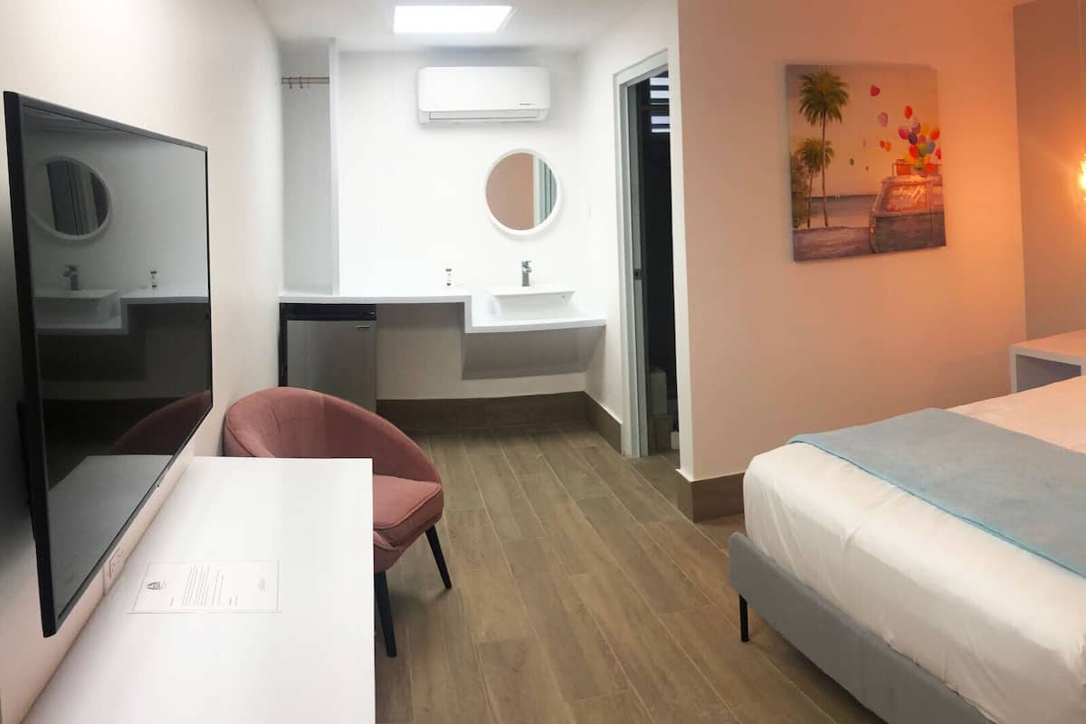 A hotel room with a bed, bedside tables, a desk with a chair, a mirror, a sink, and a painting on the wall.