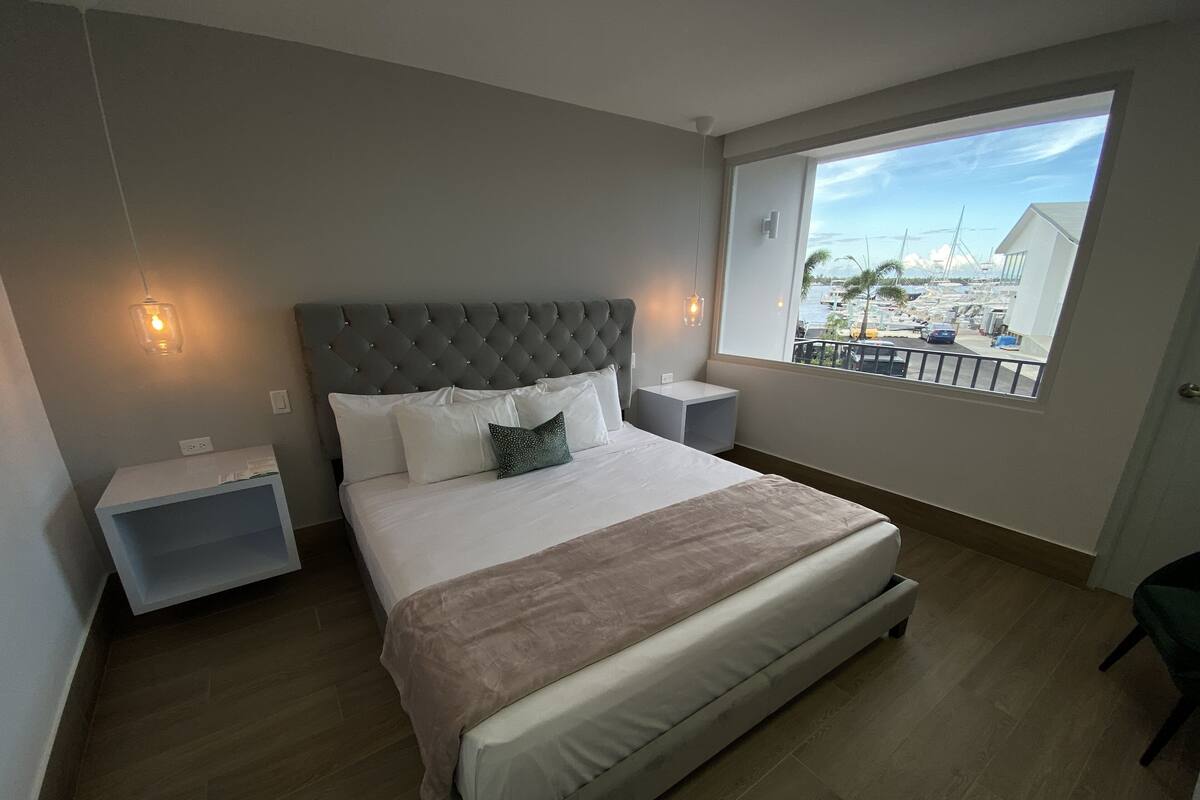 A modern bedroom with a large bed, two bedside tables, a window with a view of a marina, and a grey tufted headboard.