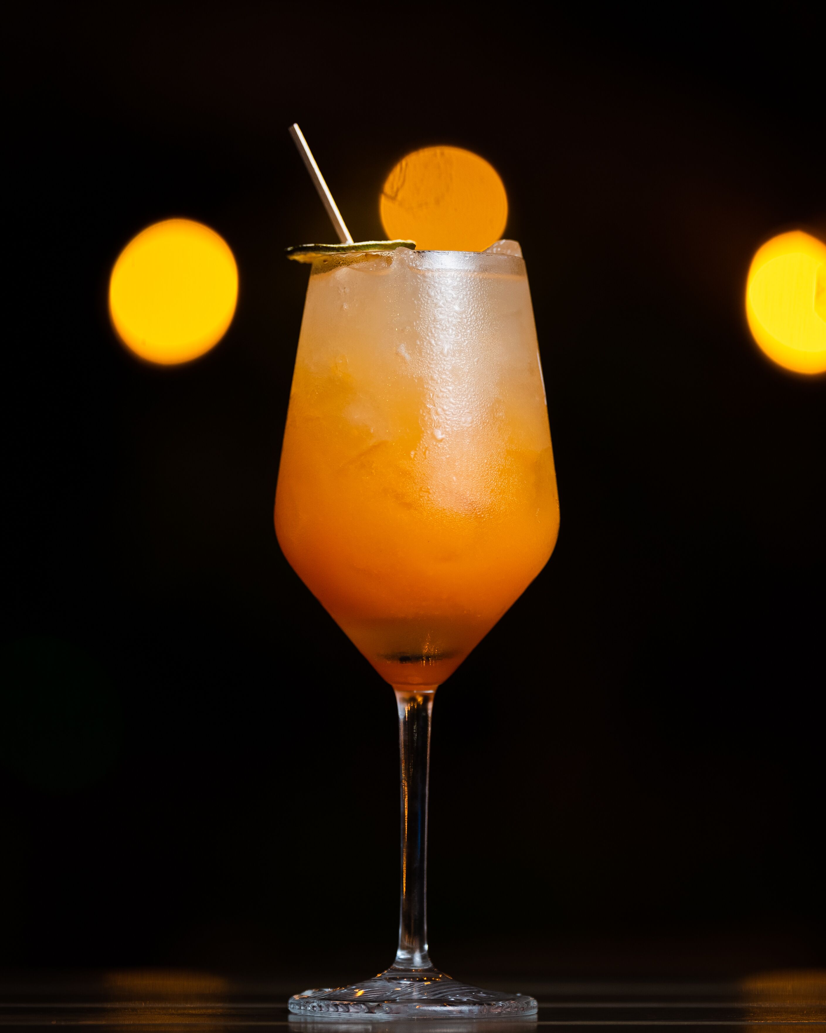 Een cocktail in een martini-glas, versierd met een citroen en een rietje, voor een donkere achtergrond met twee vage lichtjes.