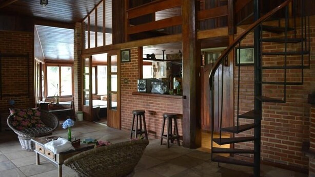 A spacious living area with a brick fireplace, a wooden staircase, and a dining area with a brick wall and large windows.