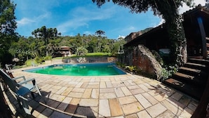 A swimming pool with a stone deck, surrounded by greenery and a rustic building.