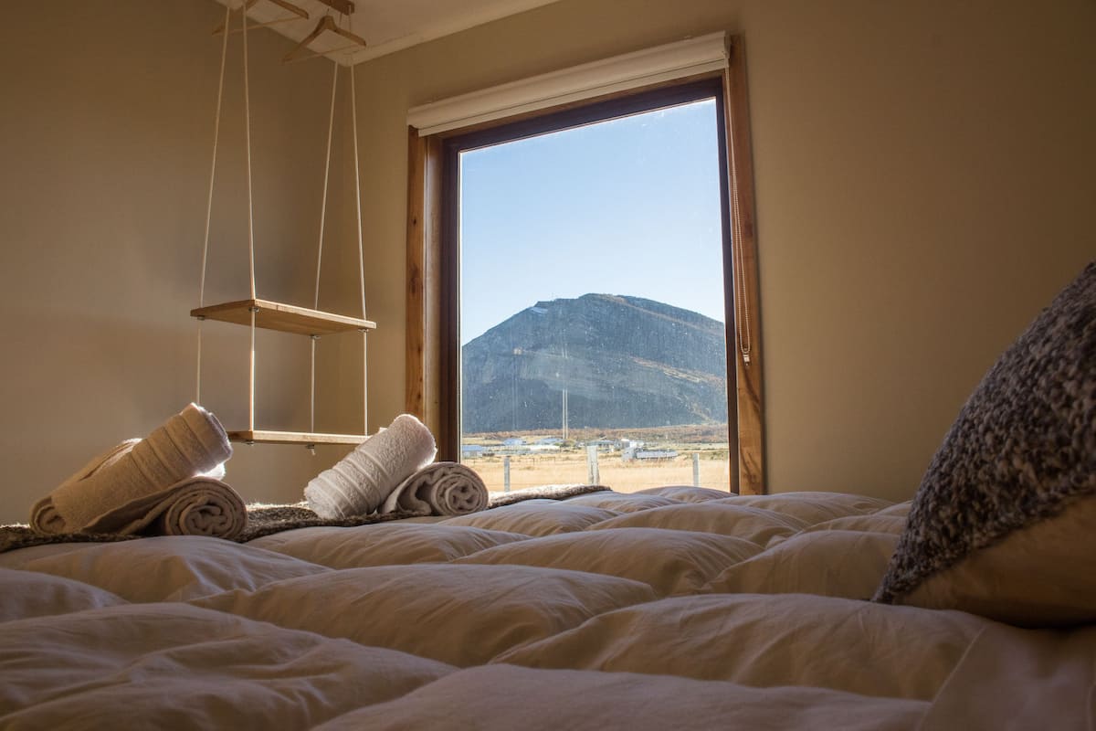 Quarto com cama, prateleira suspensa e vista para uma montanha pela janela.