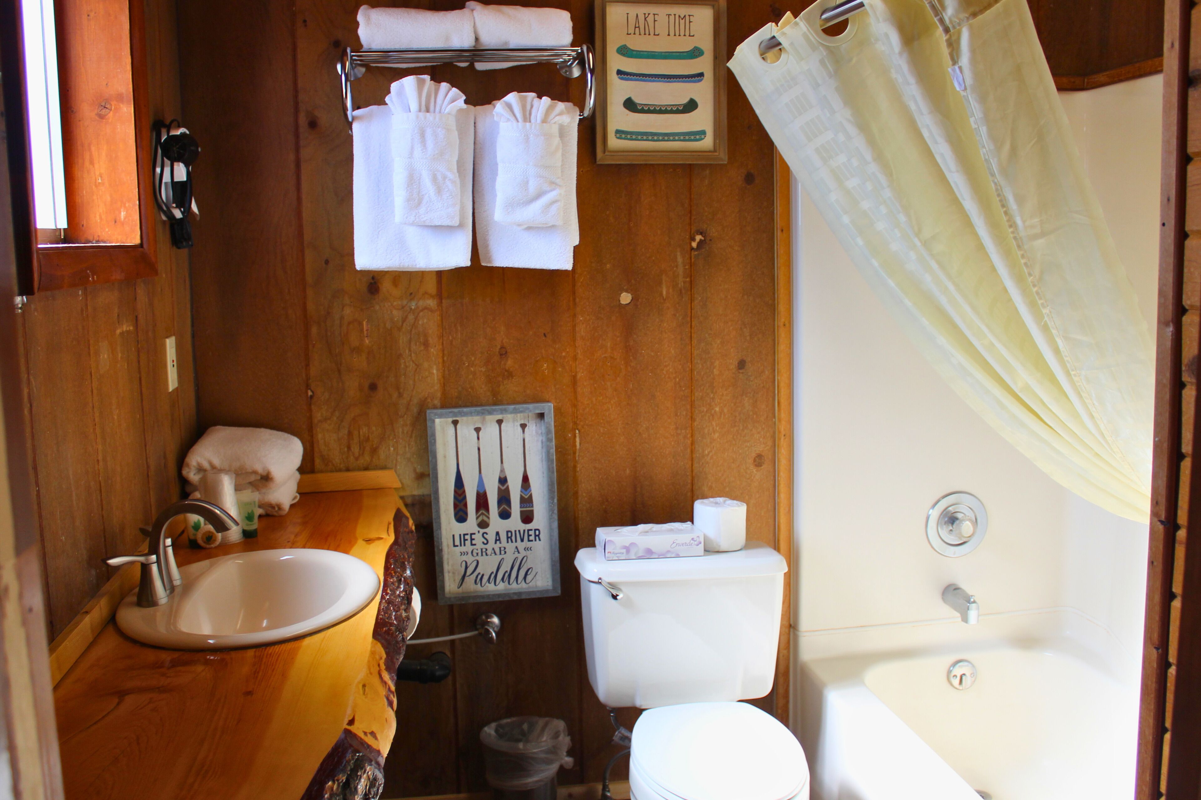 A wooden bathroom with a sink, toilet, and bathtub. There are towels hanging on a rack and a sign that says "LIFE A RIVER".
