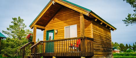 A wooden cabin with a green roof, a porch with a railing, and a single door.