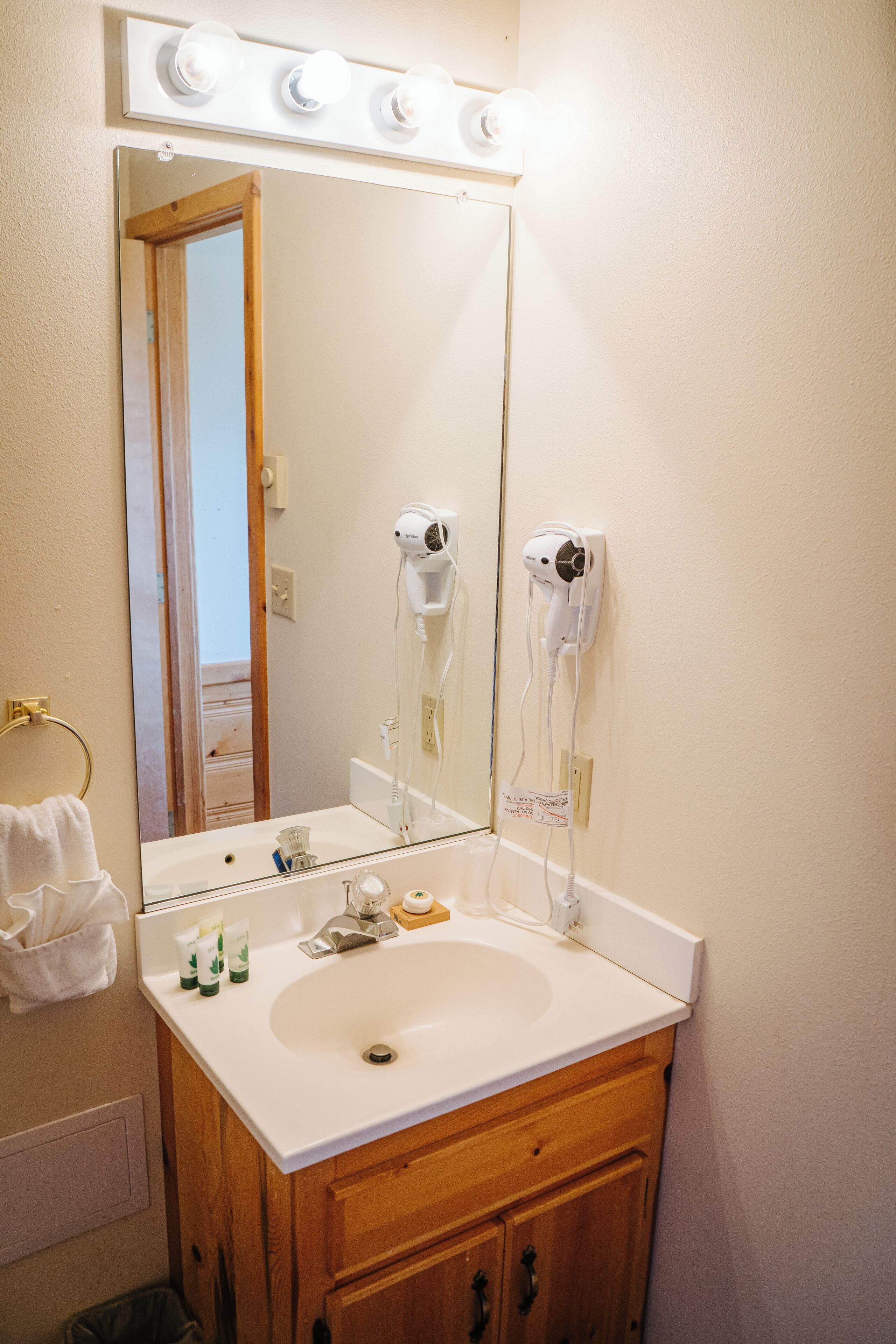 A bathroom with a white sink, wooden cabinets, a large mirror, and two hair dryers mounted on the wall.