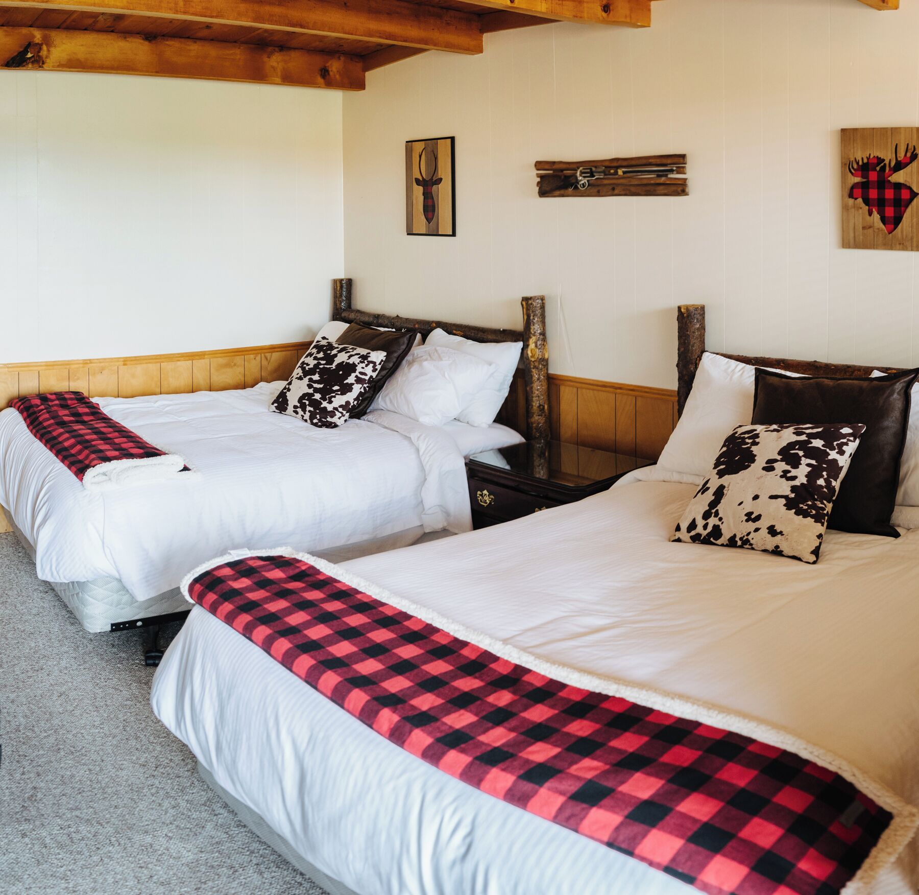 A hotel room with two beds, each with white bedding and a red and black checkered throw blanket. There are wooden headboards and a wooden beam ceiling.