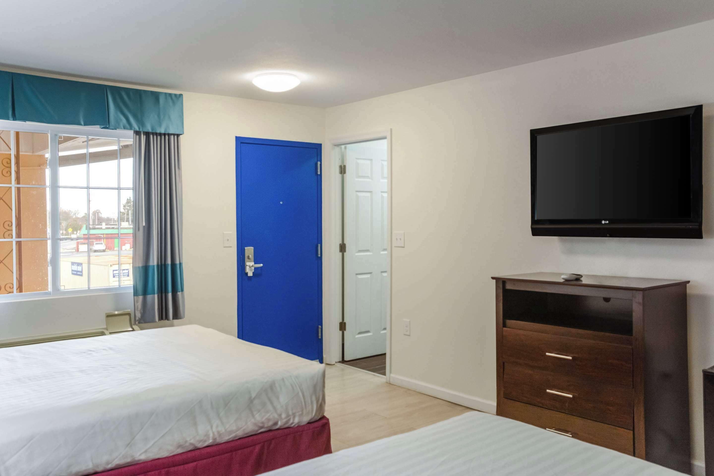 A hotel room with two beds, a television, a wooden dresser, and a blue door.