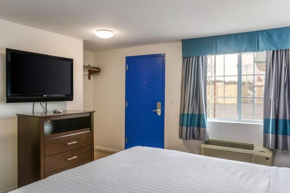 A hotel room with a bed, a flat-screen TV, a blue door, and a window with curtains.