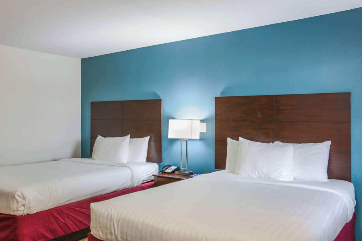 A hotel room with two beds, a nightstand with a lamp, and a blue accent wall.