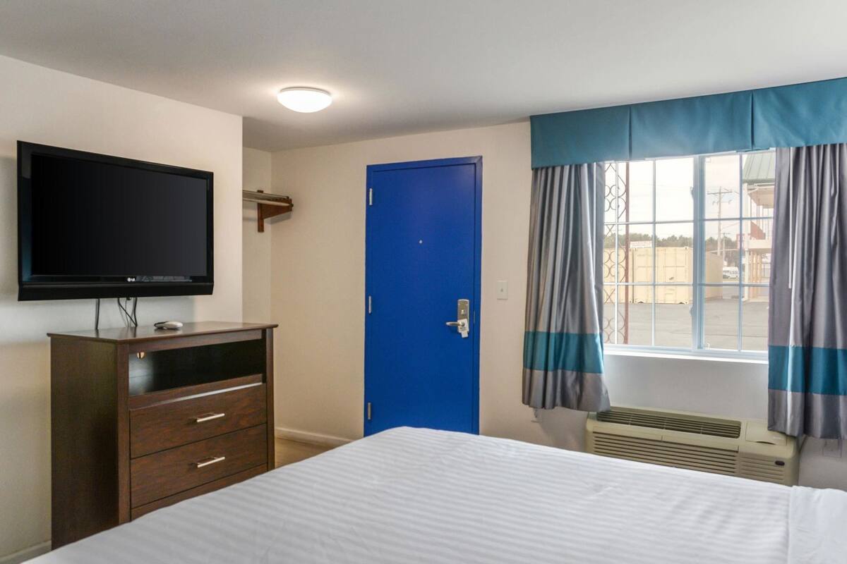 A hotel room with a bed, a TV mounted on the wall, a blue door, and a window with curtains.
