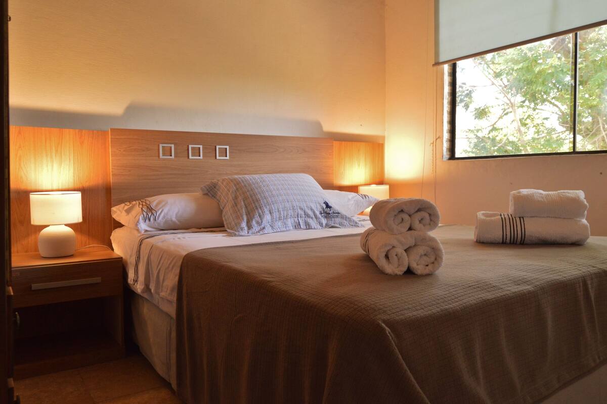 A neatly made bed with pillows, a bedside lamp, and rolled towels on a bed with a window view.
