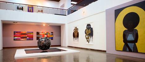 An art gallery with a central sculpture, abstract paintings, and a balcony with railings.