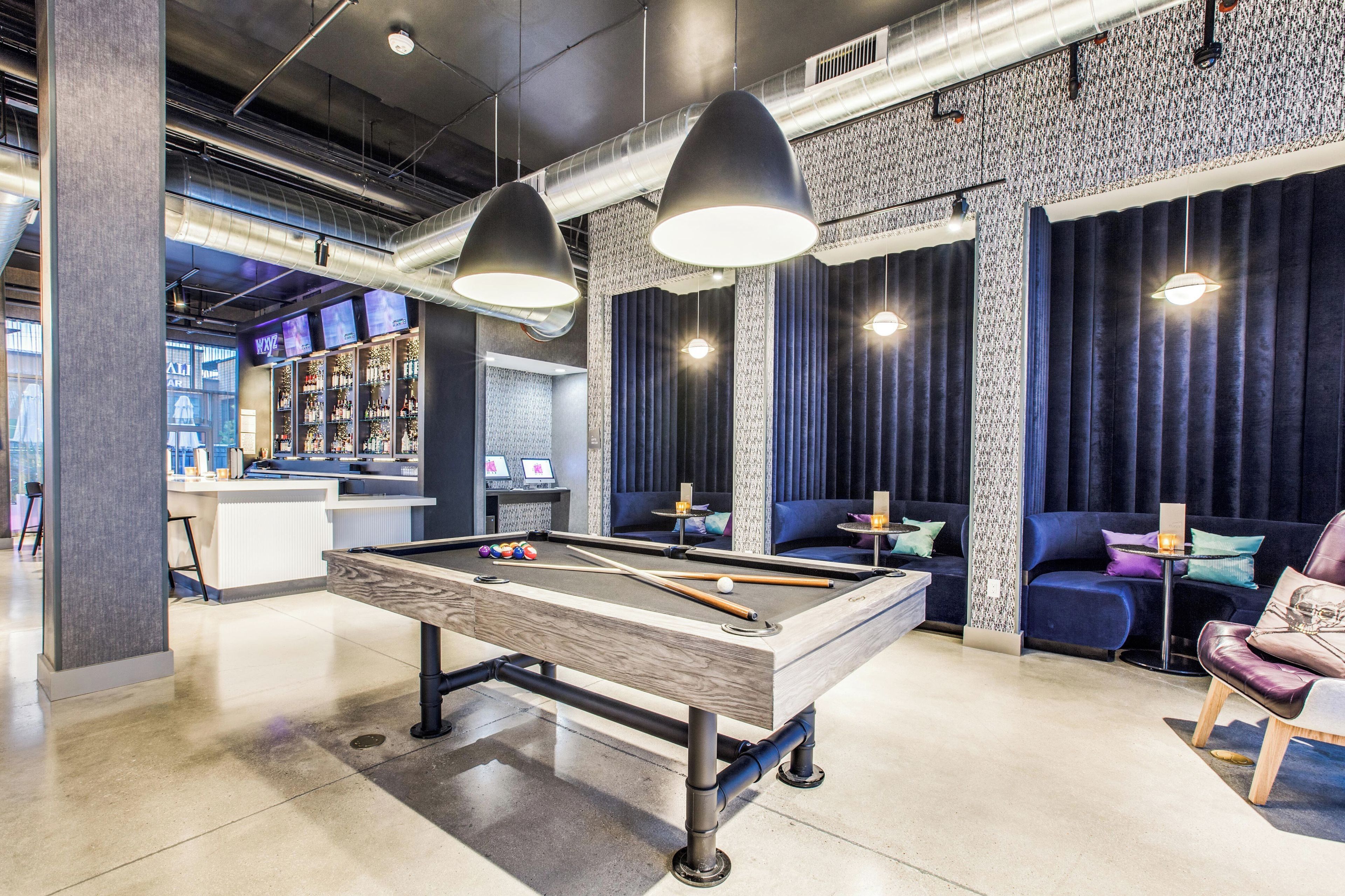 A pool table in a modern lounge area with overhead lighting and a bar in the background.