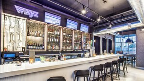 A bar area with a neon sign, multiple shelves of bottles, a counter with a digital screen, and bar stools. - Aloft by Marriott Dallas Arlington Entertainment District (Arlington)