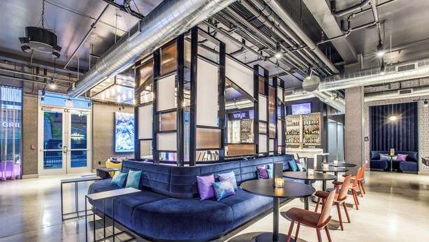 A modern lounge area with a curved blue sofa, a round table, and orange chairs. - Aloft by Marriott Dallas Arlington Entertainment District (Arlington)