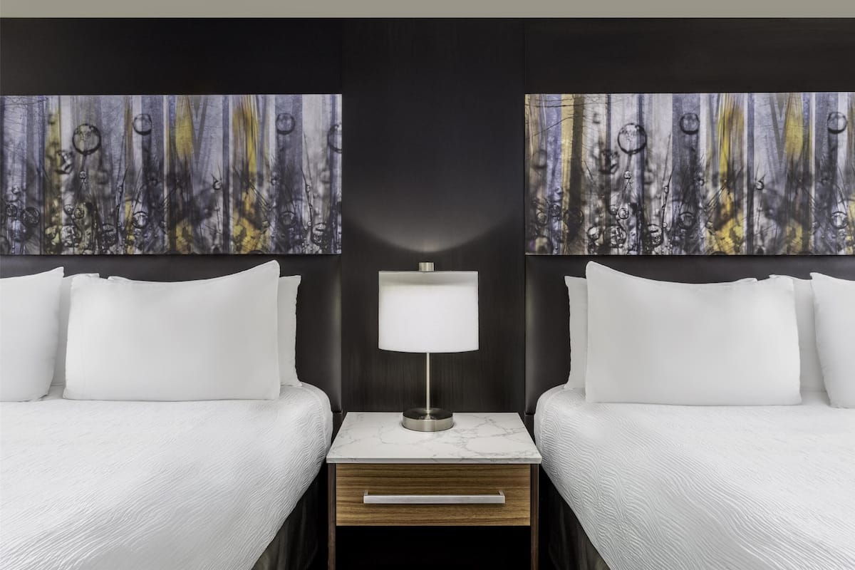 Ein Hotelzimmer mit zwei Betten, einem Nachttisch mit Lampe sowie zwei abstrakten Bildern an den Wänden.