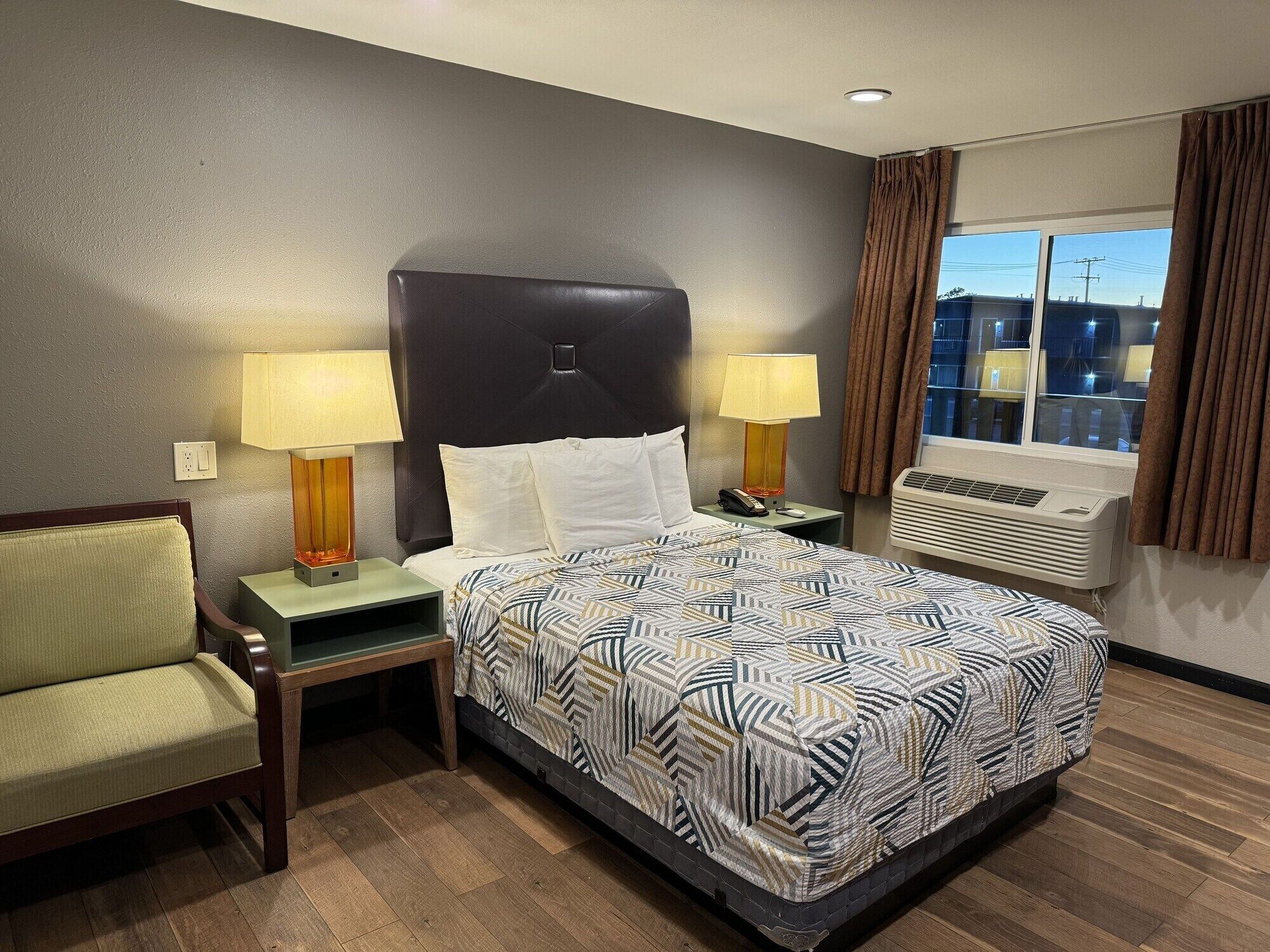 Una camera d'hotel con un letto, due lampade da comodino, una sedia, un tavolino notturno, una finestra con tende e un condizionatore.
